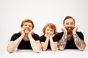 happy boy with redhead father and bearded tattooed grandpa lying and smiling at camera on light grey background