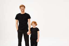 joyful redhead father and son in black t-shirts and trousers holding hands and smiling at camera isolated on grey