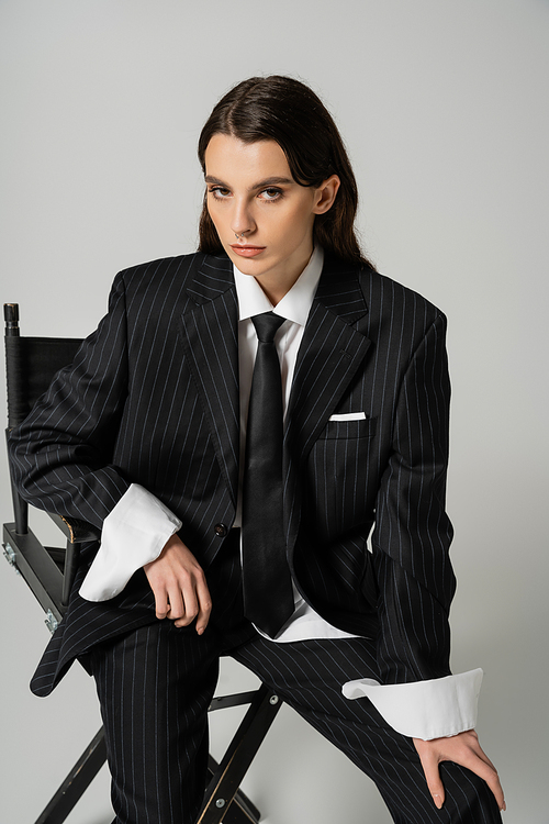 stylish brunette woman in black striped suit and tie posing on chair and looking at camera isolated on grey