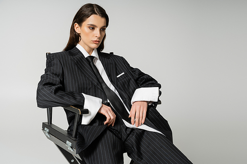 pretty brunette woman in white shirt and black striped suit sitting isolated on grey