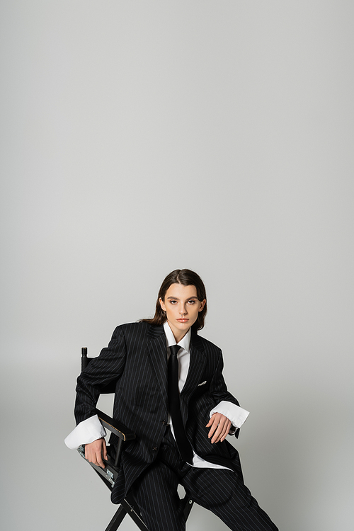 trendy woman in oversize formal wear sitting and looking at camera isolated on grey with copy space