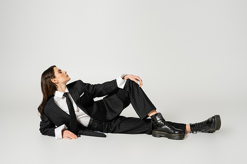 full length of young woman in stylish formal wear and laced-up boots lying on grey background