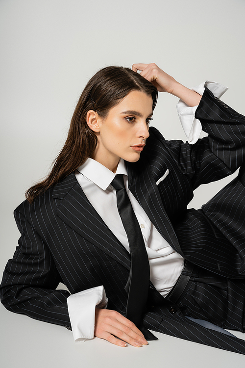 stylish woman in black striped suit and white oversize shirt posing with hand near head on grey background