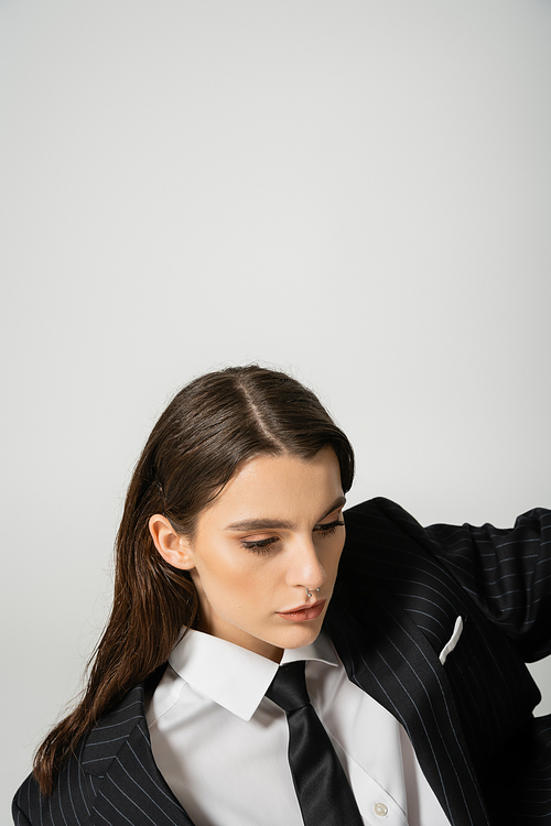 brunette woman with makeup and piercing posing in black striped suit and tie isolated on grey