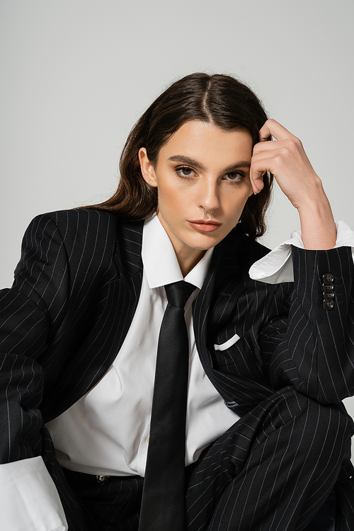 pretty woman in white shirt and black blazer with tie holding hand near head and looking at camera isolated on grey