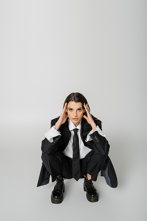 brunette woman in black striped suit and rough boots holding hands near face and looking at camera while sitting on grey