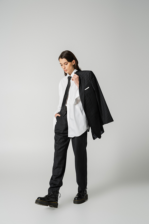 young model in oversize shirt and black pants holding black blazer while walking in rough boots on grey background