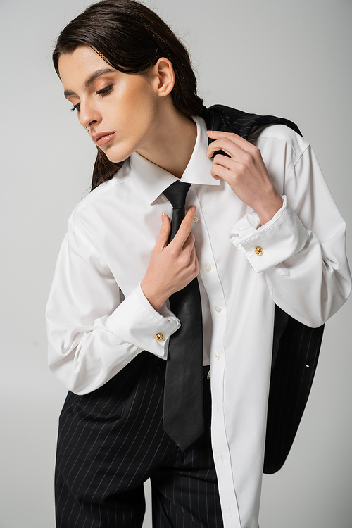 trendy woman in oversize shirt touching black tie while posing isolated on grey