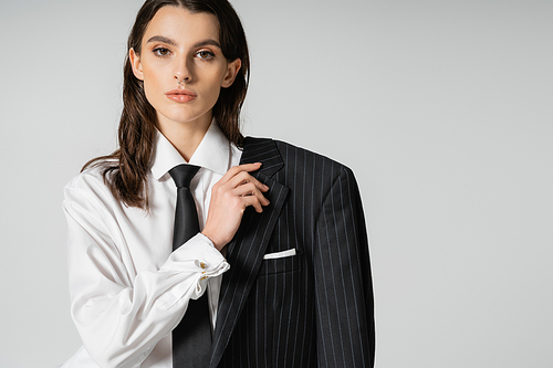 trendy woman in white shirt and tie holding black striped blazer and looking at camera isolated on grey