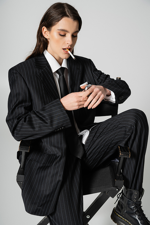 trendy woman in black oversize suit sitting with cigarette and lighter isolated on grey