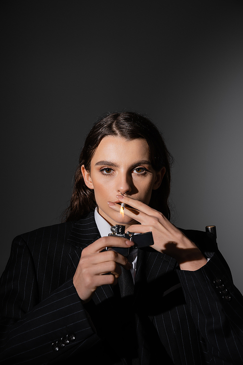 stylish brunette woman in black blazer lighting cigarette and looking at camera on dark grey background