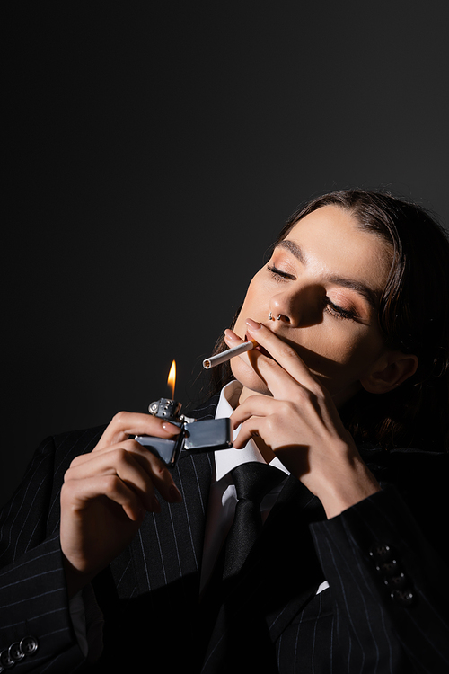 young and elegant woman in formal wear lighting cigarette isolated on black