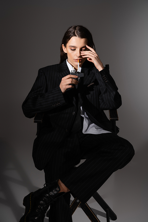 fashionable woman in black pantsuit lighting cigarette while sitting on dark grey