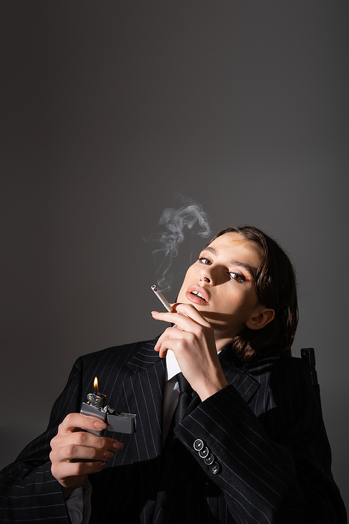 sensual woman in black striped suit holding lighter and smoking isolated on grey with copy space