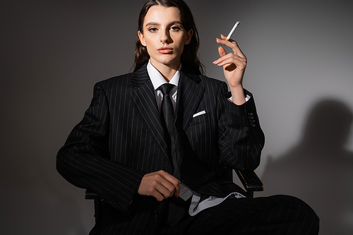 brunette woman in elegant suit sitting with cigarette and looking at camera on dark background with shadow