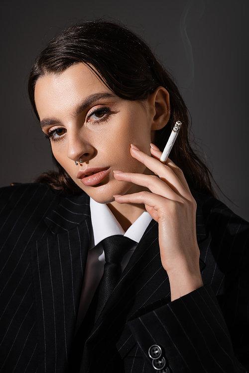 portrait of brunette woman in black striped blazer holding cigarette and looking at camera isolated on dark grey