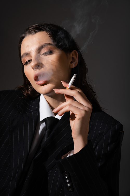 sensual brunette woman in black blazer and tie smoking isolated on dark grey