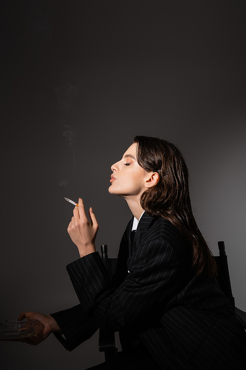 side view of brunette woman in black blazer holding ashtray and smoking on dark grey background