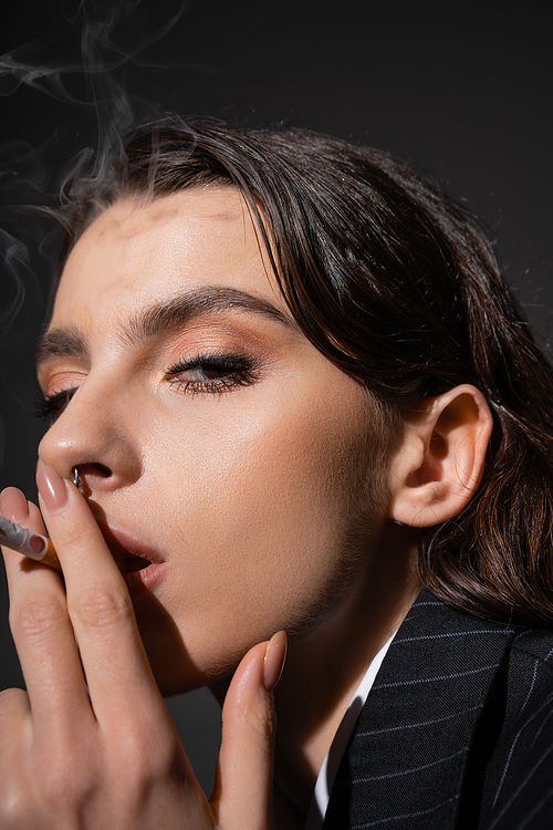 portrait of stylish brunette woman smoking and looking at camera on black background