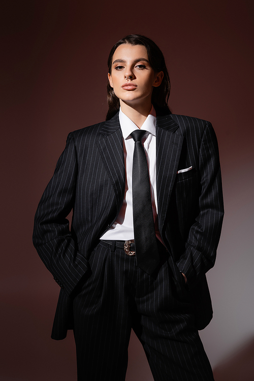 trendy woman in black striped pantsuit and tie standing with hands in pockets and looking at camera on brown background