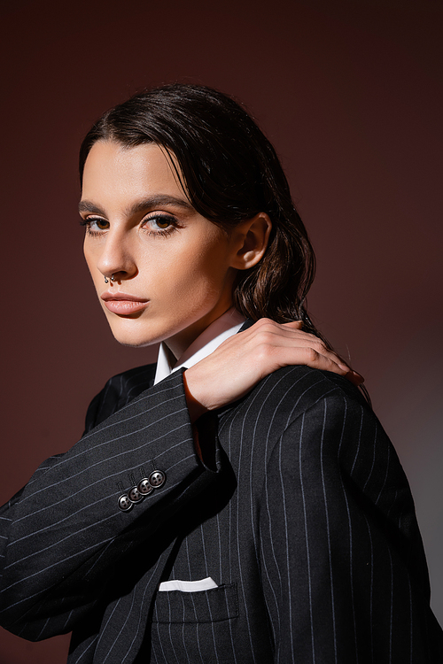 portrait of elegant brunette woman in black and striped blazer posing with hand on shoulder on brown
