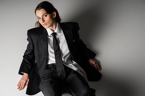 high angle view of stylish model in white shirt and black oversize suit looking at camera while sitting on grey background