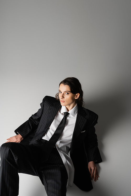 high angle view of brunette woman in black oversize suit and white shirt sitting and looking at camera on grey background