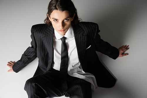 high angle view of young stylish woman in black suit and tie looking at camera while sitting on grey background