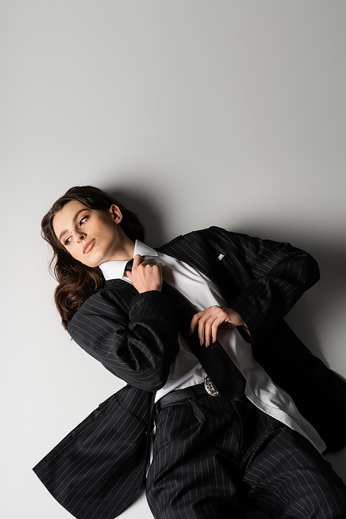 high angle view of brunette woman in black oversize suit touching tie and looking away while lying on grey background