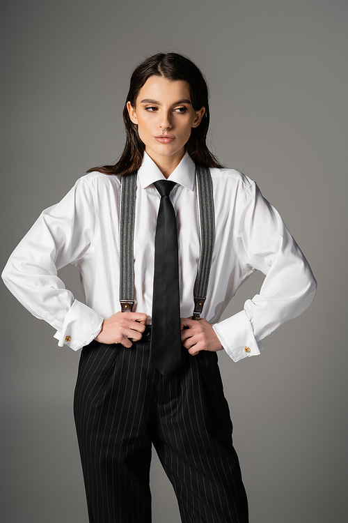 brunette woman in white oversize shirt and black tie holding black trousers with suspenders isolated on grey