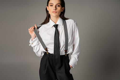 pretty brunette woman in white shirt and tie standing with hand in pocket of black trousers with suspenders on grey background
