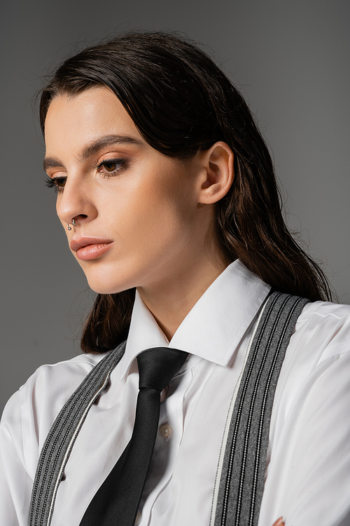 portrait of brunette woman with makeup and piercing wearing white shirt and black tie isolated on grey