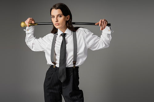 trendy woman in white shirt and black trousers with suspenders posing with walking stick and looking at camera isolated on grey