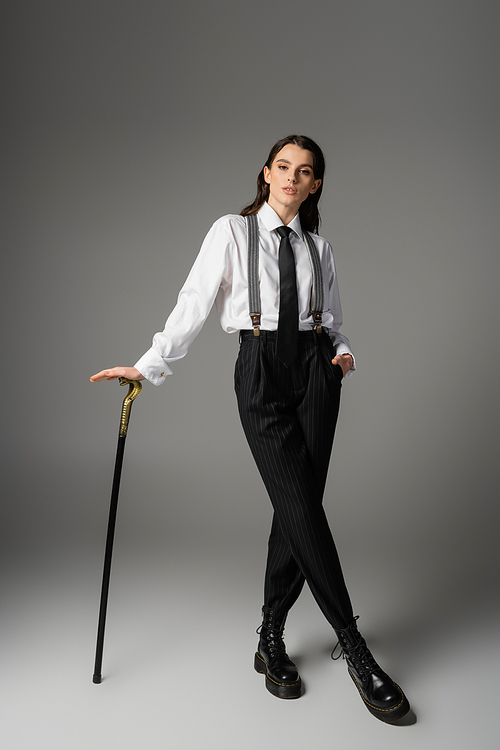 full length of young woman in elegant clothes and rough leather boots posing with walking cane on grey background