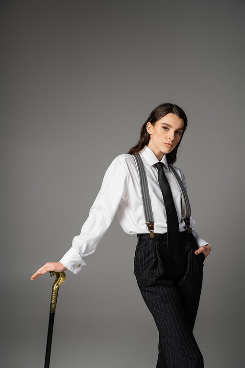 pretty brunette model in white shirt and tie posing with walking cane and hand in pocket of black pants isolated on grey