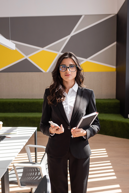 brunette businesswoman in eyeglasses and black blazer standing with smartphone and notebook in modern office