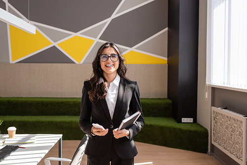 happy brunette businesswoman in eyeglasses looking at camera while standing with smartphone and notebook in office