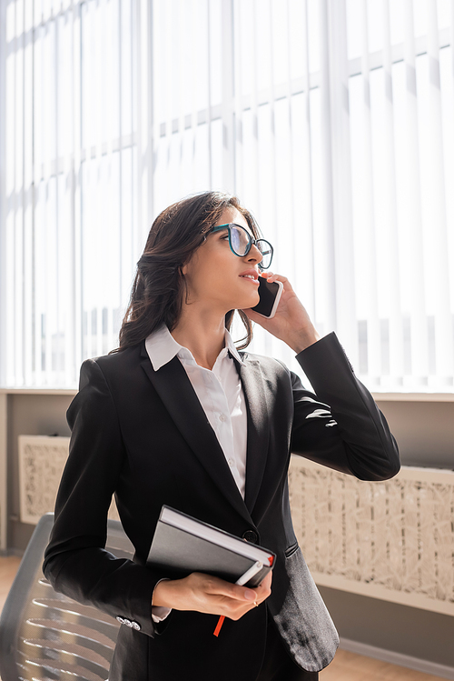 brunette office manager in eyeglasses holding notebook and looking away while talking on mobile phone