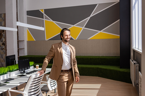 smiling businessman in beige suit standing near desk and looking away in modern office