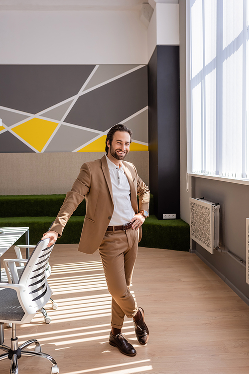 full length of happy businessman in beige suit standing with hand on hip in spacious office