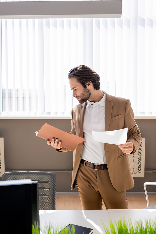 thoughtful businessman in beige suit looking at documents near desk in office