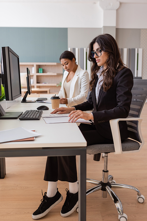 interracial businesswomen in formal wear working with documents and computers in office