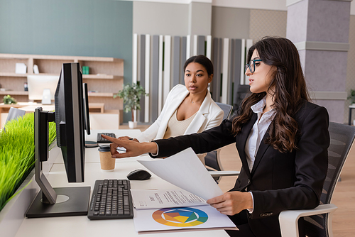 manager in eyeglasses holding paper and pointing at computer monitor near multiracial colleague