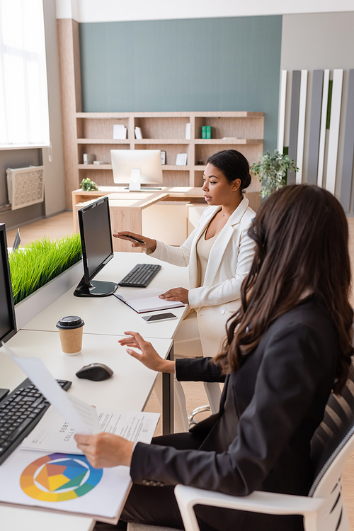 multiracial businesswoman pointing at computer monitor near colleague working with documents