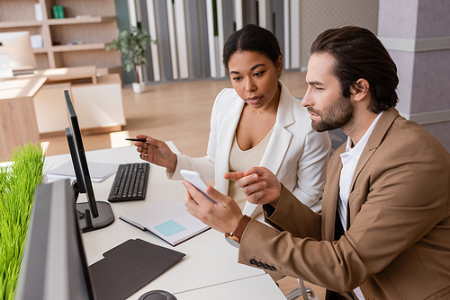 thoughtful businessman pointing at smartphone while working with multiracial colleague in office