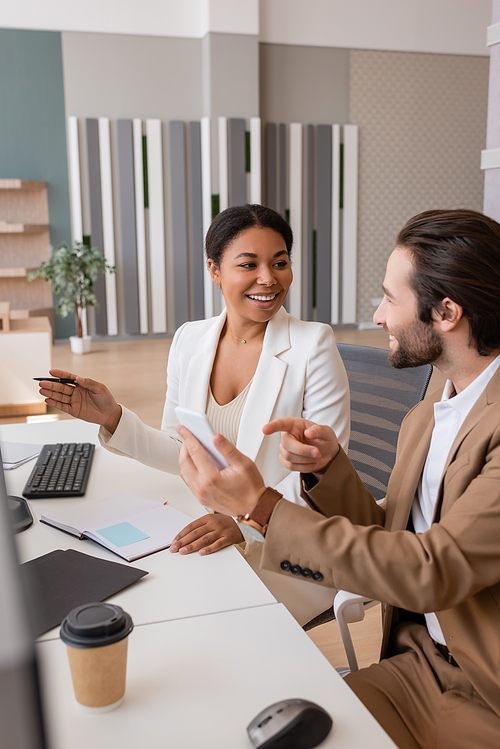 smiling manager pointing at mobile phone near multiracial businesswoman in office