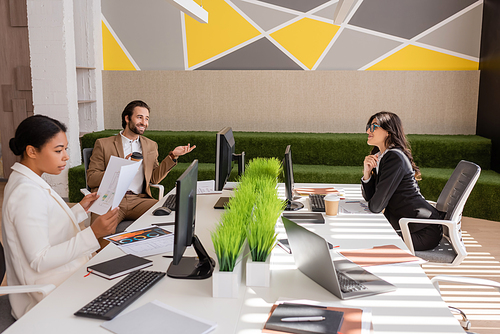 smiling businessman talking to multiethnic colleagues near computers and documents in modern office