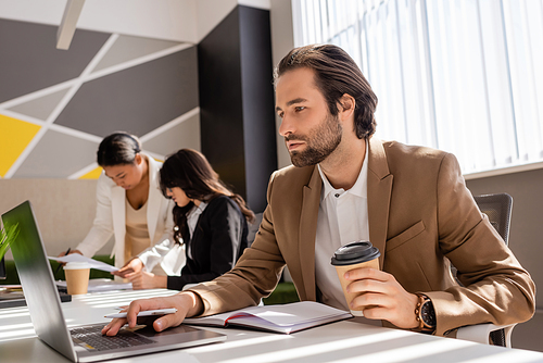 focused businessman with coffee to go using laptop near interracial colleagues working on blurred background