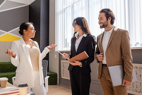 smiling managers with laptop and folder looking at positive multiracial businesswoman talking and gesturing in office