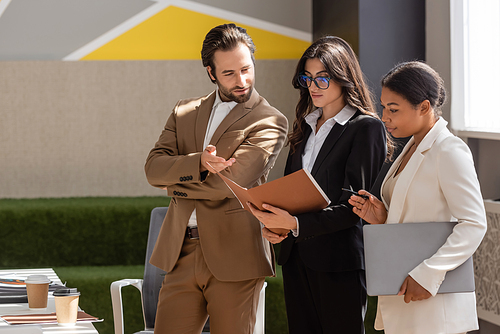 businesswoman in eyeglasses holding folder with documents near multicultural colleagues in office